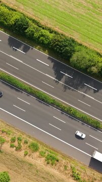Vertical screen. Aerial perspective captures vehicles moving along a highway bordered by lush greenery and fields, illustrating the continuous flow of traffic