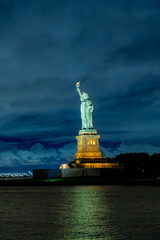 Statue of Liberty with illuminated pedestal over New York Harbor. Oblique night view across dark water, with layered clouds, tree silhouettes and distant port lights. © white78
