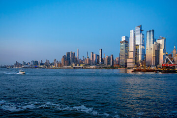 Obraz premium Midtown Manhattan skyline at twilight viewed over water in New York. Highrise modern towers illuminate evening sky above blue river water with small boat passing by.