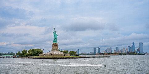 Statue of Liberty view with watercraft in New York Harbor. Monument on island surrounded by water with moving jet skis, city skyline under gray clouds, wide angle shot. © white78