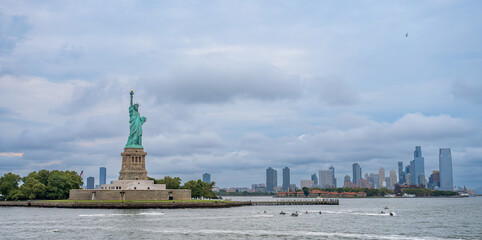 Statue of Liberty on Liberty Island in New York Harbor. Green copper monument on stone pedestal near water with jet skis, city skyline under overcast sky, wide shot. © white78