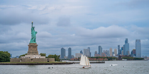 Statue of Liberty and sailing ship in New York Harbor. Green copper statue on stone pedestal near white schooner, city skyscrapers under overcast blue sky, side view. © white78