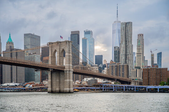 Brooklyn Bridge across East River with Lower Manhattan skyscrapers background. Stone towers with steel cables stretch over water surface leading to urban financial center under cloudy sky.