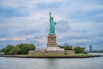 Statue of Liberty monument on Liberty Island in New York. Green statue on stone pedestal surrounded by trees, water foreground under cloudy blue sky, medium shot. © white78