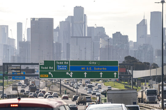 Busy commuter traffic on the West Gate Freeway (M1) approaching the Todd Road exit and service center with the Melbourne city skyline visible in the background. Infrastructure, city connectivity.