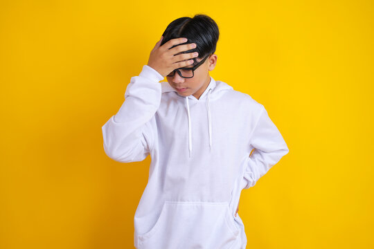 Stressed Asian Teenage Boy Wearing Glasses and White Hoodie Holding Hand on Forehead