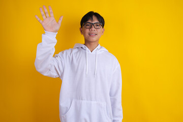 Asian Teenage Boy Wearing Glasses and White Hoodie Waving Hand Saying Hello © Gatot