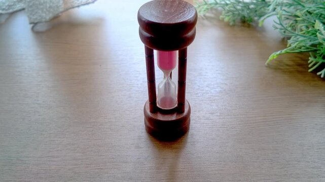 A wooden hourglass with pink sand stands on a wood table beside green foliage, symbolizing time and deadlines.