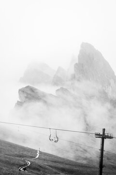 Dramatic black and white minimal fine art vertical layout empty chairlift ski lift mechanism thick mist covered Seceda mountain peaks background Dolomites Italy