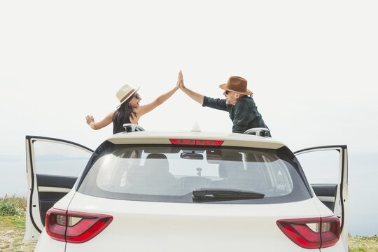 Couple cheers with arms raised Hi-Five clam hands while standing by white car overlooking coastal cliff. Bright horizon and open doors evoke sense of freedom and successful road trip journey