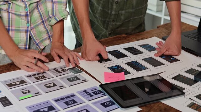 Creative web developers designing user interface layouts for a mobile application, collaborating on a new project for a smartphone screen with printed templates on a desk