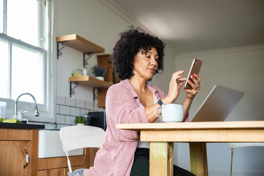 Woman using smartphone and laptop at home kitchen table