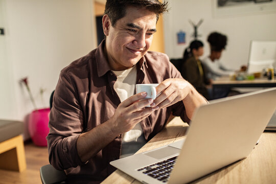 Smiling man drinking coffee while working on laptop in modern office