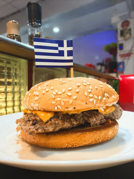 Close up of a juicy beef cheeseburger with melted cheese on a sesame seed bun topped with a miniature Greek flag toothpick
