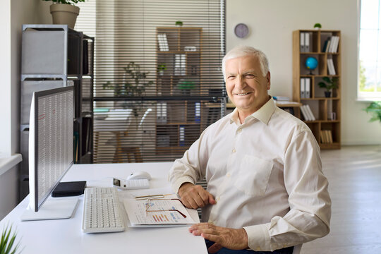 Senior man accountant at office computer workstation. He reviews analysis data and financial documents, preparing a report at a tidy desk. Clear professional finance and retirement concept.