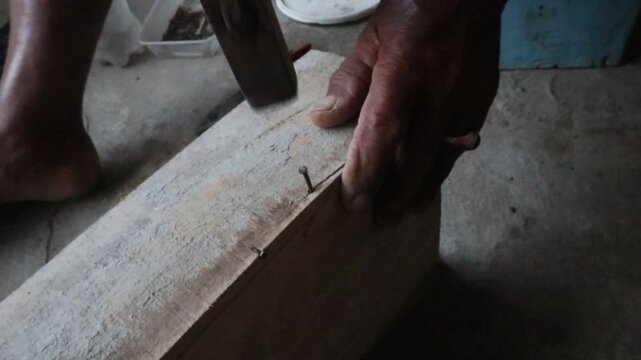 Hands using a hammer to drive a nail into a wooden board, showing traditional manual carpentry work and a practical woodworking construction process.