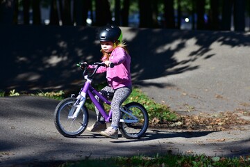 Fototapeta premium Young girl wearing a safety helmet riding a bicycle on a pump track in a sunny green park, practicing cycling skills and enjoying active outdoor recreation, exercise and childhood adventure.