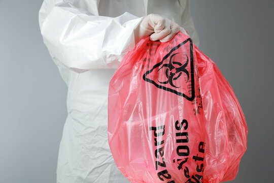 Cautious worker wearing white protective suit holds red biohazard bag full of clinical waste carefully during hazardous medical cleanup