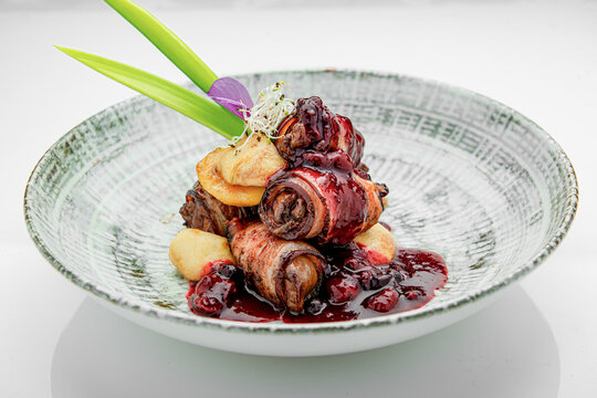 Culinary dish with meat rolls and sauce served in a decorative bowl. A plate holds a stack of meat rolls topped with dark sauce. Green garnish adds color.
