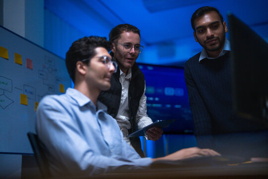 Three male specialists from different backgrounds studying the same dashboard exchange short remarks while tracking an incident across systems in a night shift operations room