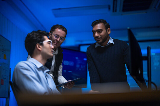 Three male engineers from diverse backgrounds solving a problem at one desk exchange short remarks while tracking an incident across systems in a night shift operations room