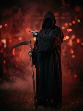 Halloween grim reaper costume featuring skull mask and black tattered robe holding wooden scythe in smoky room with blurred crowd party background.