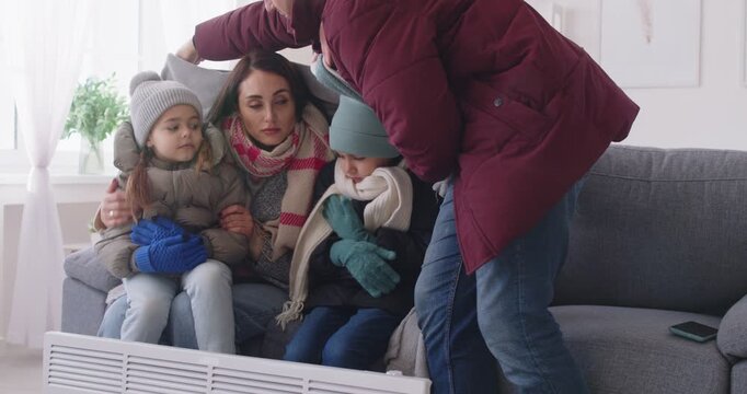 Caring father covering with plaid his wife and children suffering from low temperature. Freezing young family sitting on sofa near electric heater, hugging and trying to warm up during cold winter.
