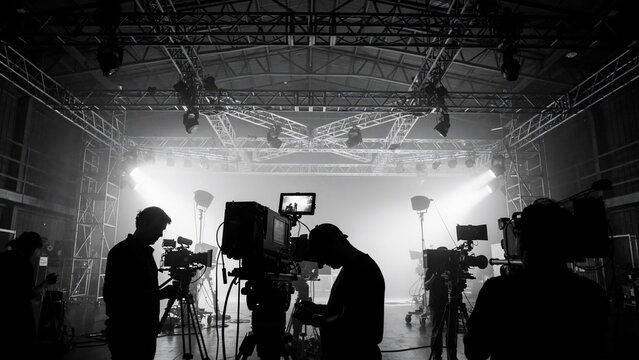 Dramatic black and white scene of silhouetted film crew with professional video cameras under bright stage lights and truss, concept for film production, TV broadcasting and media content
