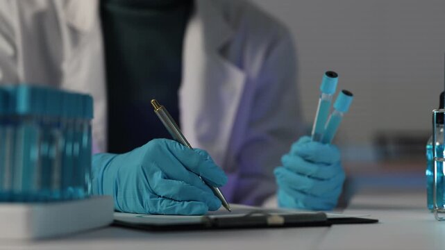 Scientist in a lab coat and blue gloves writing notes on a clipboard and holding test tubes with blue liquid, researching a new coronavirus vaccine or developing a drug
