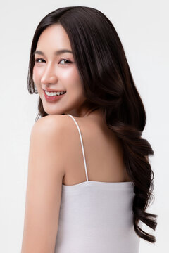 Beauty portrait of a smiling young Asian woman looking over her shoulder with glowing skin and long wavy hair against a white background, ideal for haircare and cosmetics.