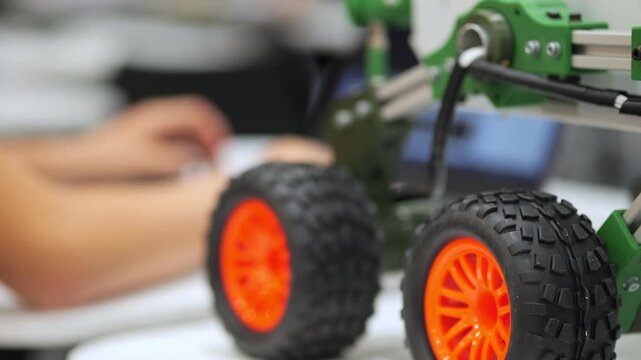 Close-up of green robotic vehicle with orange wheels. Hands work on device in background. Focus on mechanical parts and tire details