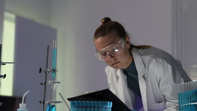 Focused female chemist in lab coat and safety goggles performing a titration experiment and recording data on a clipboard in a modern scientific research laboratory