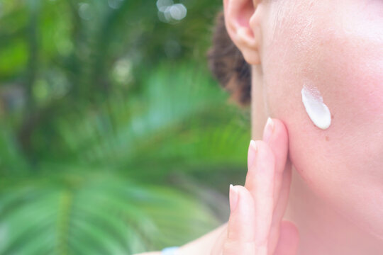 Young woman applying moisturizer to her face against green palm leaves.
