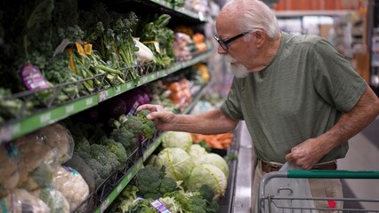 An elderly man is selecting fresh vegetables at a grocery store. He is focused on choosing the best...