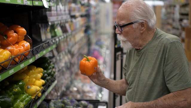 Senior man is shopping in a grocery store and looking at colorful bell peppers in the produce section while making choices