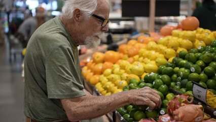 An elderly man examines fruits in a grocery store. He stands in front of a display filled with colorful citrus and tropical fruits © Robert Peak