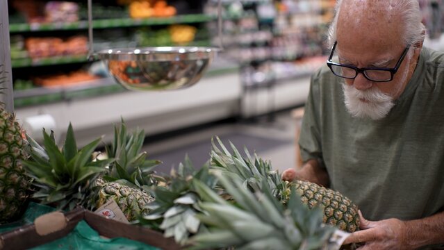 An elderly man inspects fresh pineapples while shopping in a grocery store during the daytime. He focuses on quality and freshness