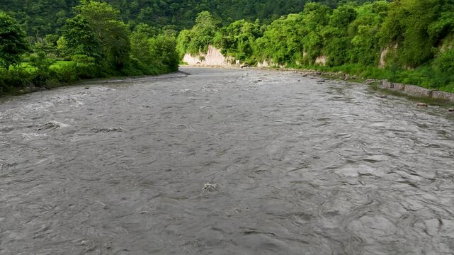 Low angle view of a wide muddy river rushing rapidly through a dense green forested valley in the Himalayan foothills of Nepal