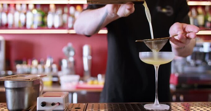 Pouring cocktail. Cold alcohol drink. Professional male barkeeper straining yellow aperitif using metal sieve preparing on counter.