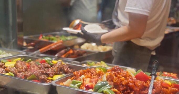 Worker in black gloves serving white rice at Chinese food buffet. Assorted stir-fry dishes in metal trays at restaurant food stall. Asian takeaway service background.