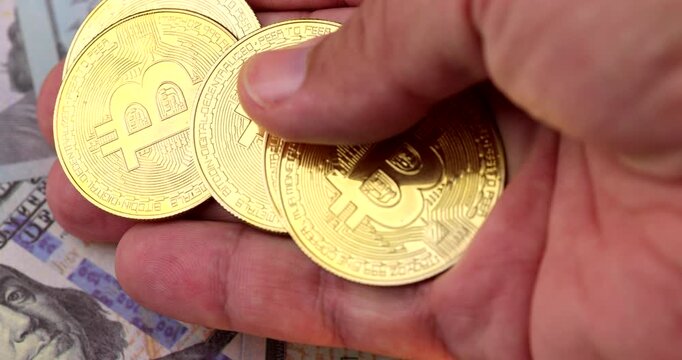 Horizontal macro view of hand arranging shiny golden bitcoin coins into a neat grid pattern over spread US hundred dollar paper banknotes.