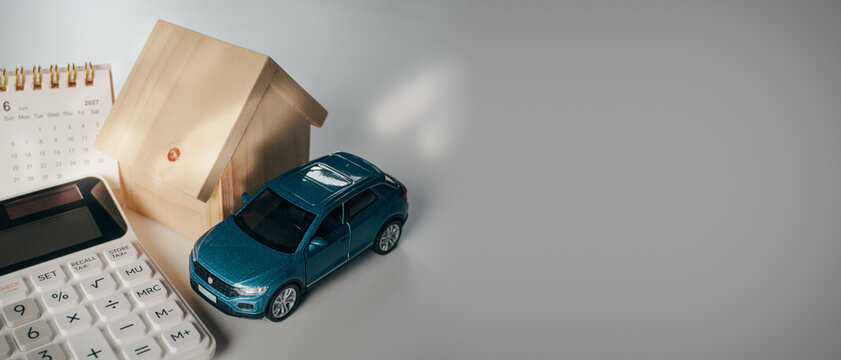 Miniature car, wooden house model, June 2027 calendar, and calculator on white desk with copy space, concept of financial planning, loan for car and house purchase, and asset investment.