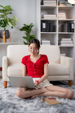 Young woman working on laptop at home sitting on carpet with books and bookshelf behind