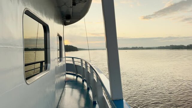 A cruise ship deck overlooks the calm river at sunrise. Golden sunlight reflects off the water and windows. The serene scene evokes a feeling of peaceful tranquility.
