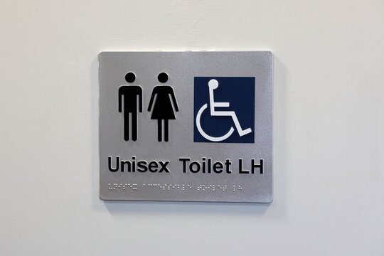 Silver restroom sign mounted on a white wall, featuring male and female pictograms next to a blue wheelchair accessibility icon, with text reading "Unisex Toilet LH" and braille lettering
