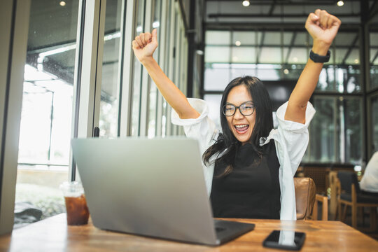 Happy woman celebrating a successful achievement while working on a laptop in a modern cafe, representing business success, productivity, career growth, accomplishment and professional achievement.