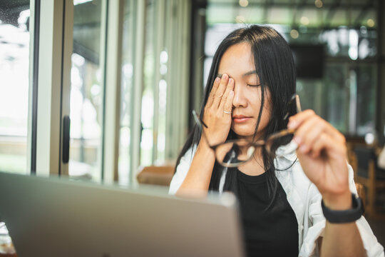 Tired woman removing glasses and rubbing her eyes while working on a laptop, illustrating eye strain, visual fatigue, prolonged screen time, stress and digital work.