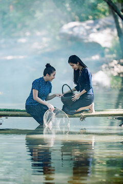 Two happy Asian women in traditional indigo dress catching fish on a bamboo bridge in Sakon Nakhon, Thailand. Perfect for travel banners and culture ads.