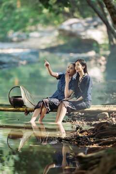 Two happy Asian women in traditional indigo dress catching fish on a bamboo bridge in Sakon Nakhon, Thailand. Perfect for travel banners and culture ads.