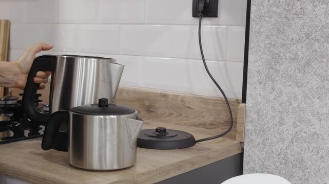 Hand unplugging stainless steel kettle from black power outlet on white tile wall in modern kitchen setting. With original sound captured.